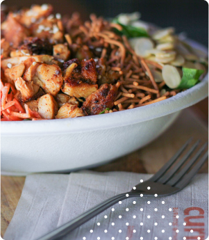 A bowl of grilled chicken salad with noodles, carrots, and vegetables sits on a table next to a fork on a napkin.